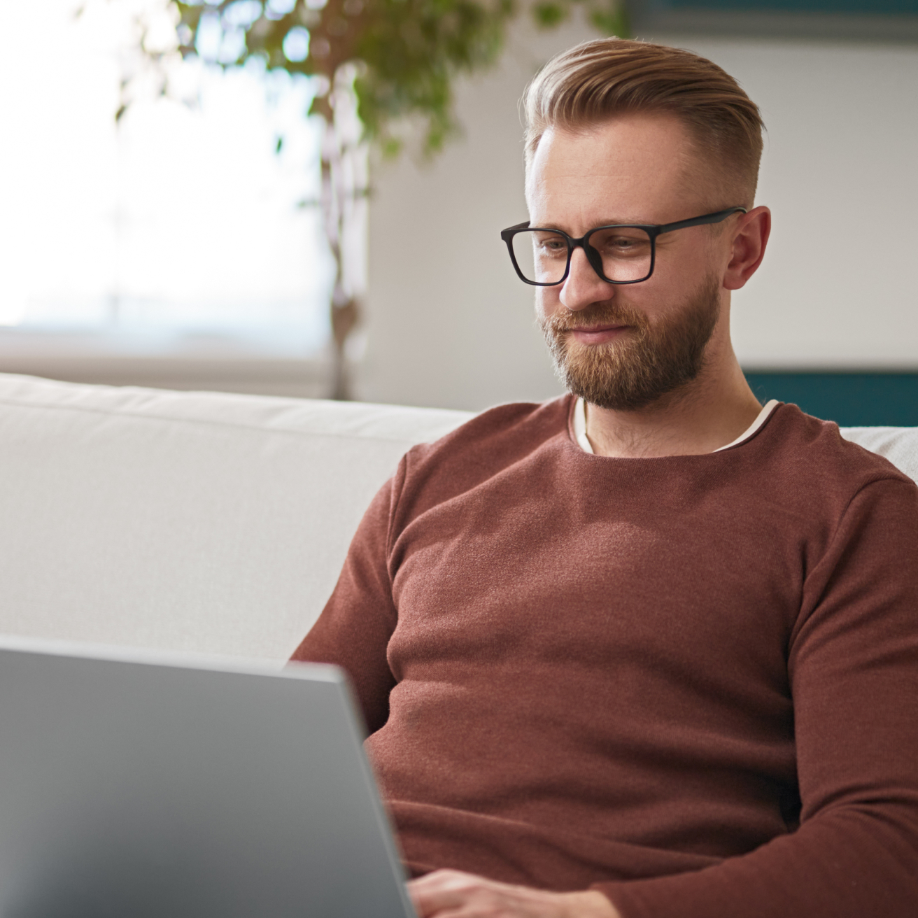 Mann mit Brille und Bart sitzt auf Sofa und arbeitet konzentriert am Laptop